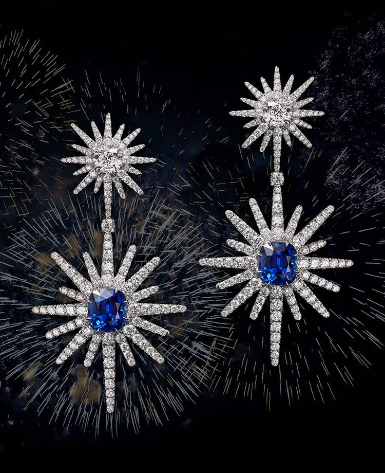 A collage shows a pair of David Yurman High Jewelry Starburst drop earrings floating in front of fireworks. The jewelry is crafted from 18K white gold with diamonds and blue sapphire center stones.