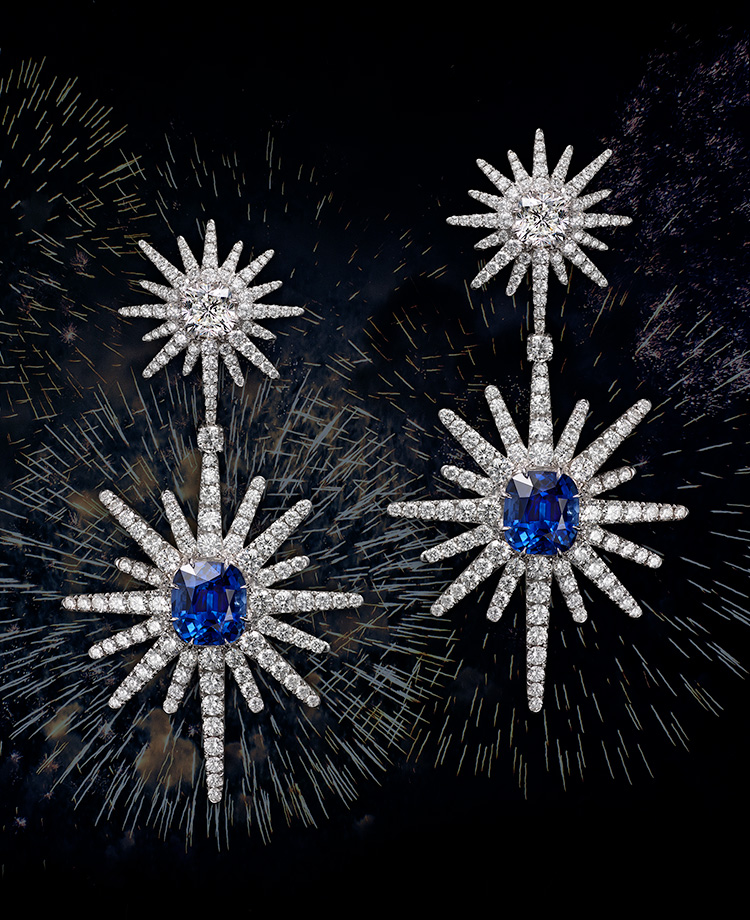 A collage shows a pair of David Yurman High Jewelry Starburst drop earrings floating in front of fireworks. The jewelry is crafted from 18K white gold with diamonds and blue sapphire center stones.