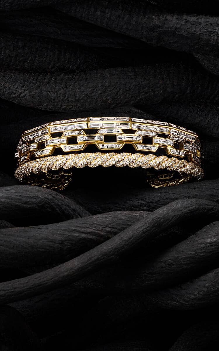 A colour photo shows a David Yurman High Jewelry Stax cuff bracelet placed in between black-hued vines. The three-row cuff bracelet is crafted from 18K yellow gold encrusted with pavé and baguette white diamonds, and combines a row of faceted metal with a row of oval link chain and a row of Cable.