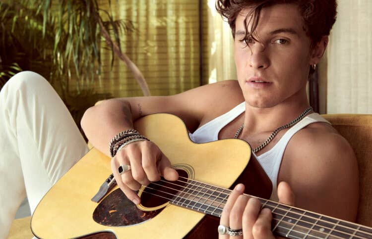 Shawn Mendes laying on a couch playing a guitar and wearing David Yurman jewellery.